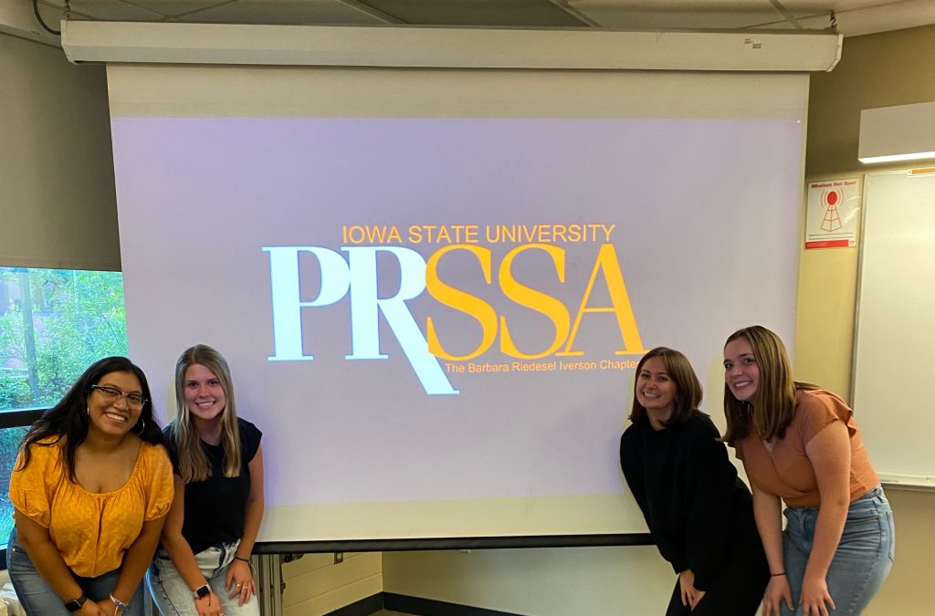 Four students smile in front of a projector screen with the PRSSA logo on it.